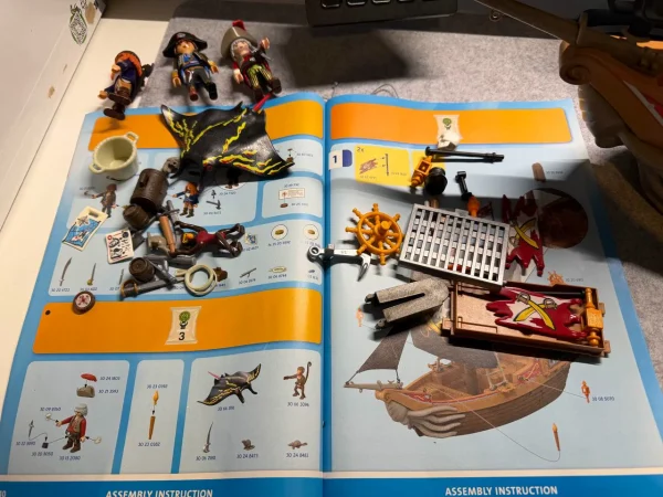  Lose Teile des Playmobil-Piratenschiffs auf der Bauanleitung, darunter Segel, Steuerrad und Anker; darüber liegen drei Piratenfiguren.