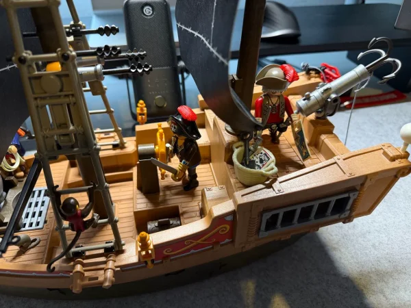 Playmobil-Piratenschiff schräg von oben mit schwarzen Segeln, Piratenfiguren, großer grauer Kanone, Fässern und zwei Ratten auf dem Deck.